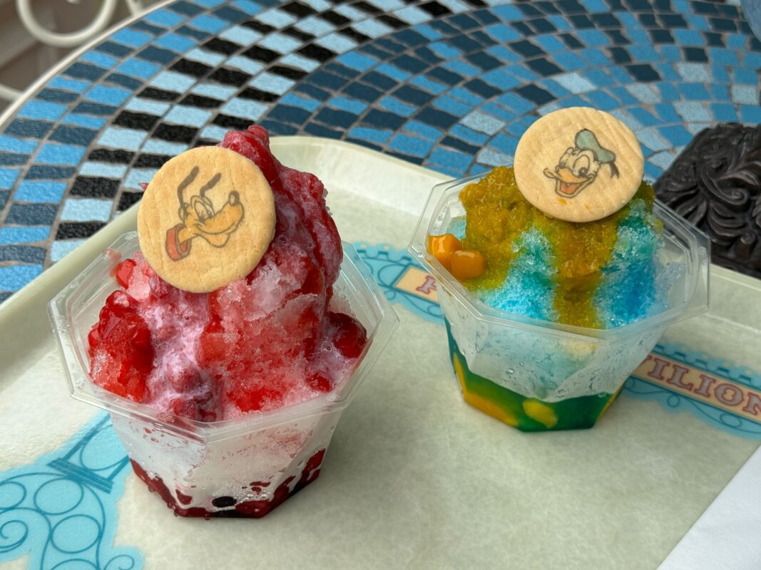 Two cups of shaved ice: the left cup features red syrup and a thin character cookie on top, while the right cup has blue and yellow syrup with a different character cookie on top. They sit on a tray.