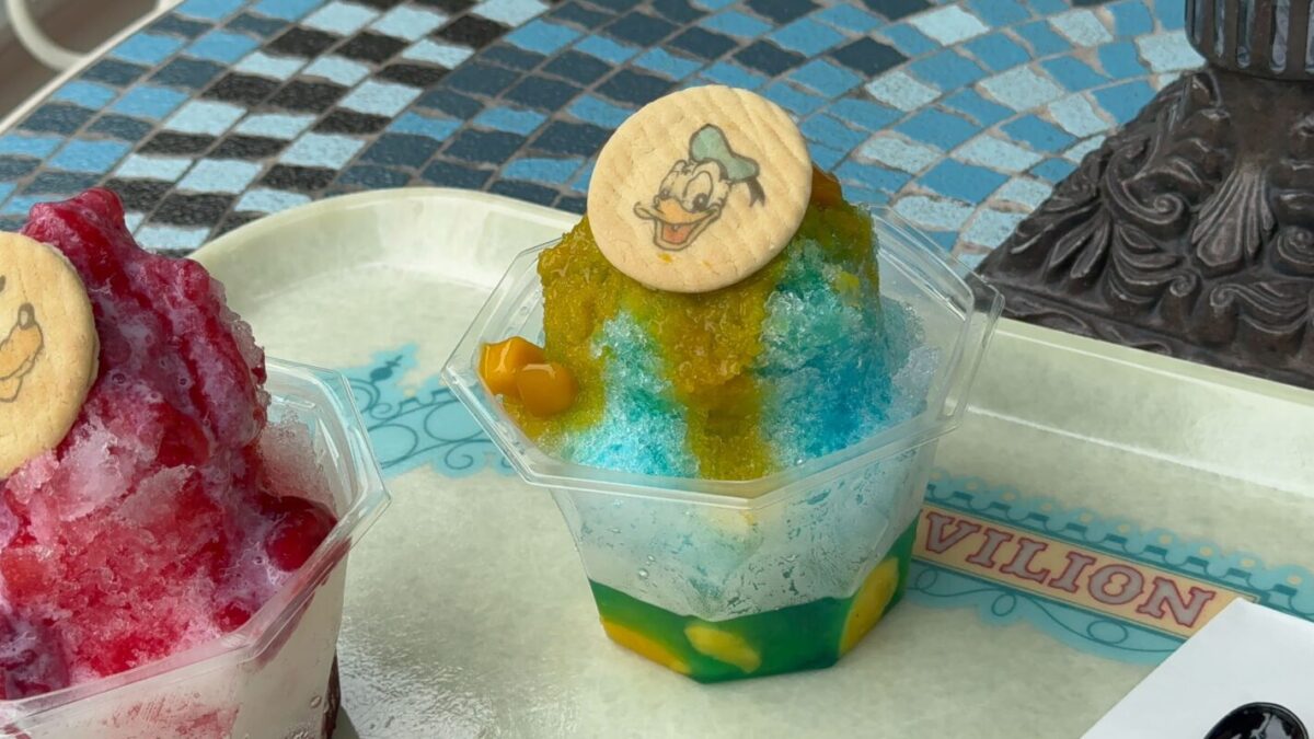 Two containers of colorful shaved ice on a tray, each topped with a character-themed cookie. The ice has different colors layered and includes small pieces of fruit at the bottom.