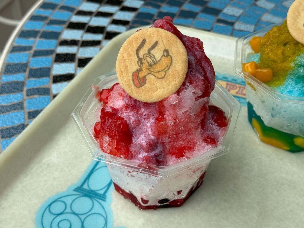 A cup of shaved ice with red syrup and a cookie featuring a cartoon dog on top, placed on a tray with a blue and black tiled background.