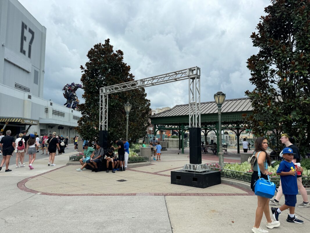 People walk and sit around a plaza with trees, metal structures, and buildings in the background under a cloudy sky.