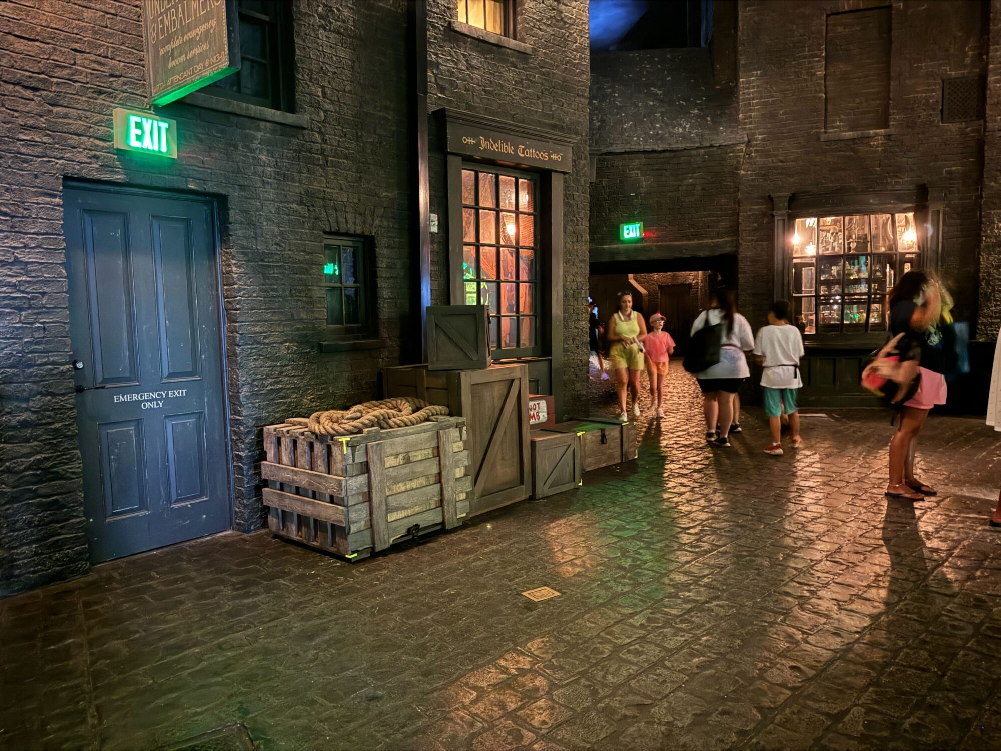 A dimly lit alley with a "Quality Quidditch Supplies" shop. A wooden crate and barrels are outside the shop. Several people are walking along the cobblestone path, passing by lit exit signs.