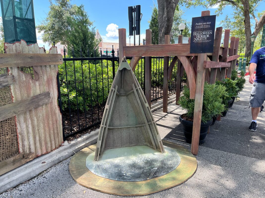 A Halloween-themed outdoor decoration setup featuring a small tent-like structure with wooden accents. Trees and plants are visible in the background. A sign references Halloween Horror Nights.