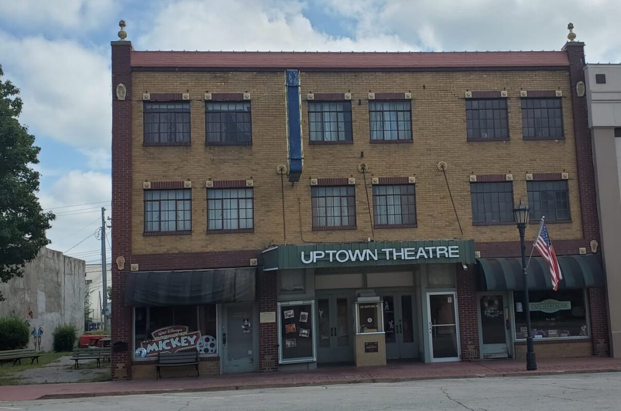 A historic three-story brick building with a sign reading "Uptown Theatre" stands proudly in the hometown. The structure features multiple windows and storefronts at street level, including a banner for "Mickey," nodding to Walt Disney. An American flag is visible on the right.