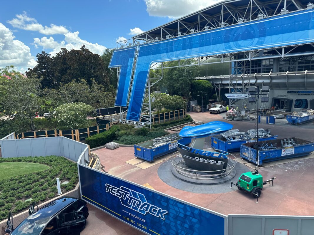 An outdoor construction site for an attraction named "Test Track" by Chevrolet, surrounded by blue fencing and equipment under a partially cloudy sky.