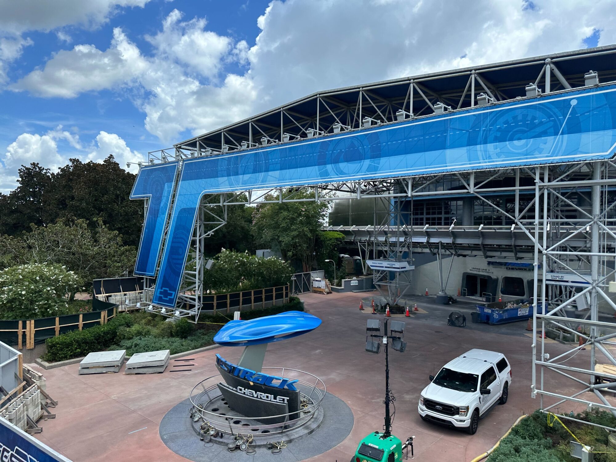 Construction site with steel framework and a blue overhang structure, featuring "Test Track Chevrolet" signage. A white vehicle is parked nearby, surrounded by construction materials and tools.