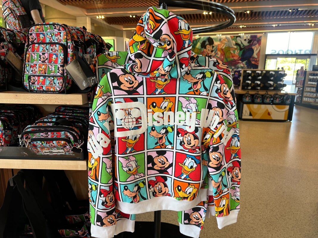 A colorful hoodie on display features a grid design with various Disney characters. Matching backpacks are also visible on the shelf. The store interior includes a window showing part of the outside.