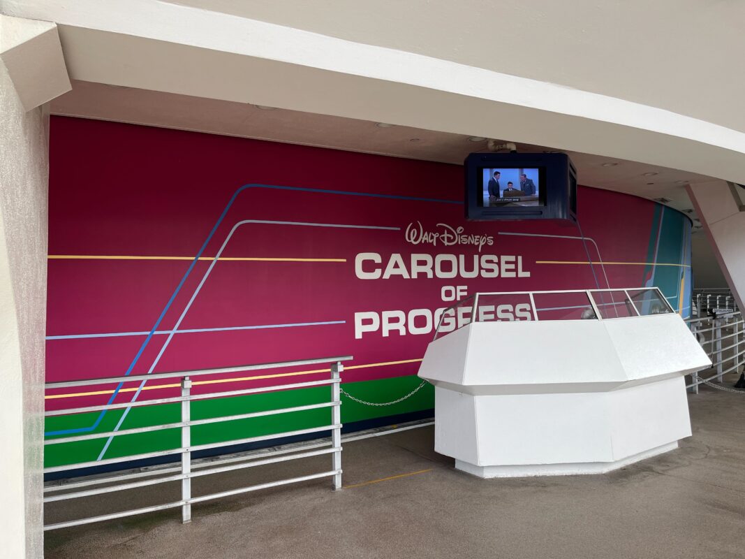 The image shows the entrance to the "Carousel of Progress" attraction at Walt Disney World, with a colorful mural and a small TV displaying information above an empty queue area.