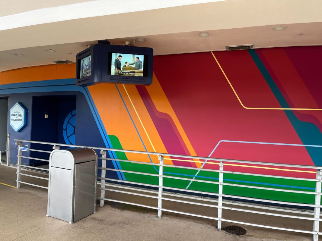 A colorful mural with diagonal lines in orange, yellow, and pink on a wall. A TV monitor above shows a person standing. A silver trash can and a railing are in the foreground.