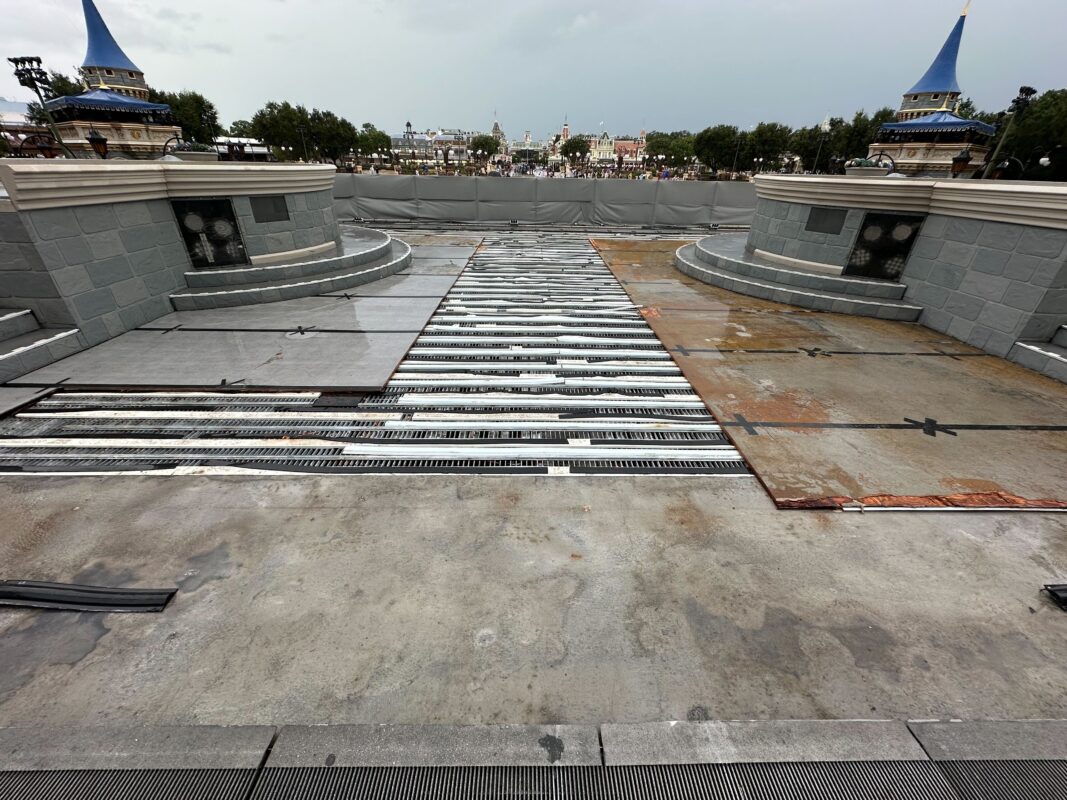 A construction site featuring a partially completed concrete surface with metal reinforcements, surrounded by gray barriers and turrets in the background.