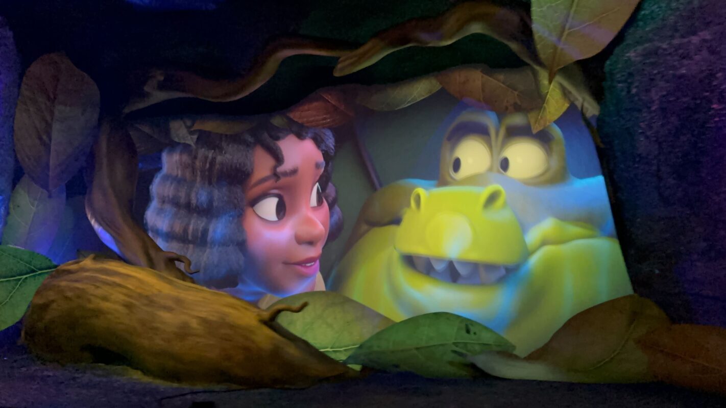 A young woman with curly hair and freckles looks closely at a large green creature with a bulbous nose and wide eyes through a forested scene.