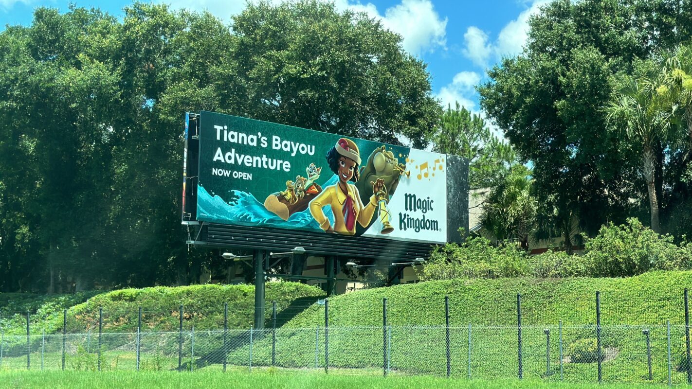 Billboard advertising "Tiana's Bayou Adventure" at Magic Kingdom featuring animated characters and a lush green background, with text reading "Now Open." Auto Draft ensures timely updates to keep the excitement fresh.