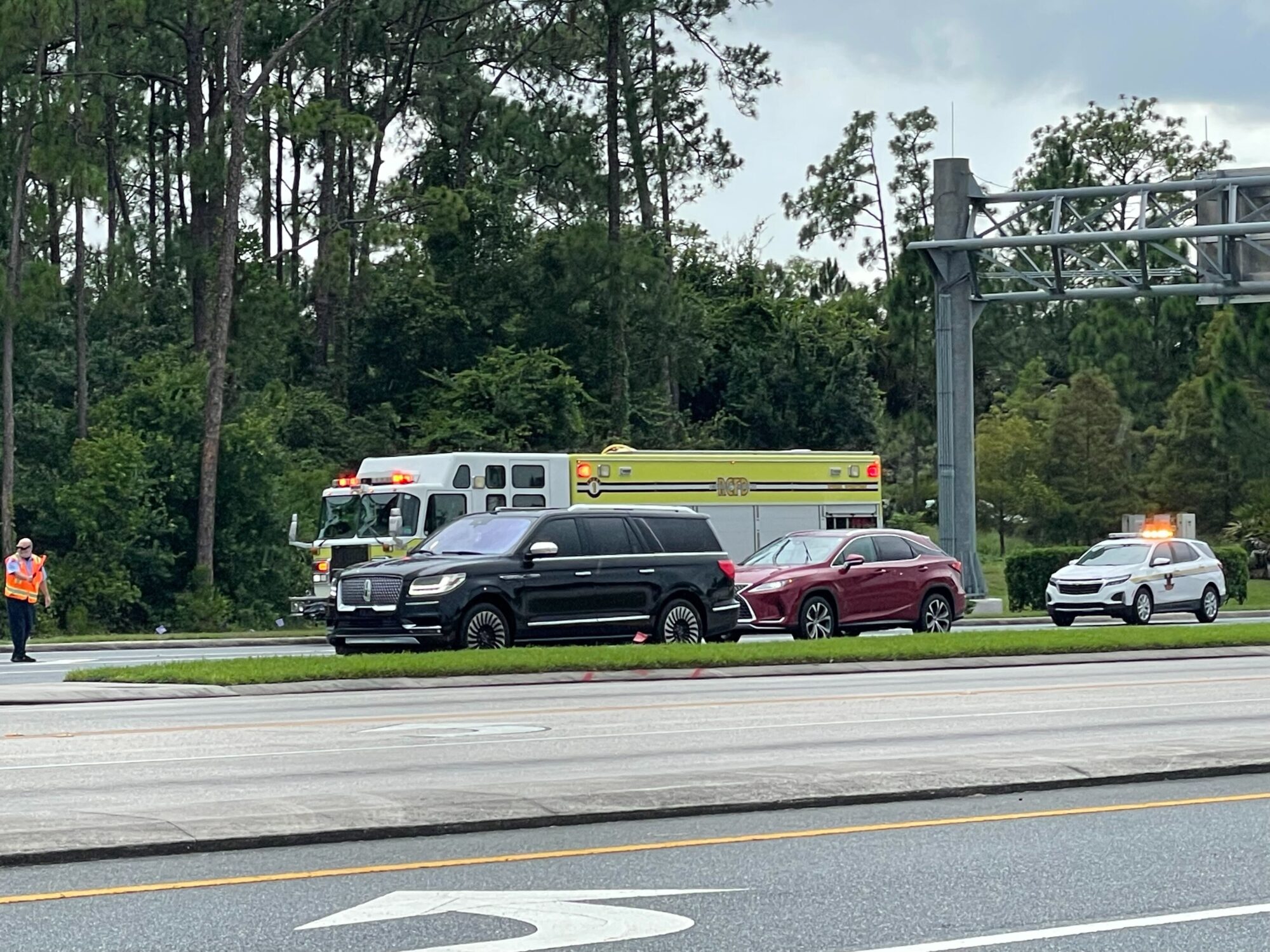 Three vehicles are parked at the side of a road, with a fire truck behind them, resembling a scene right out of Disney's Typhoon Lagoon. Two individuals in high-visibility vests stand nearby as lightning subtly illuminates the trees and greenery in the background.