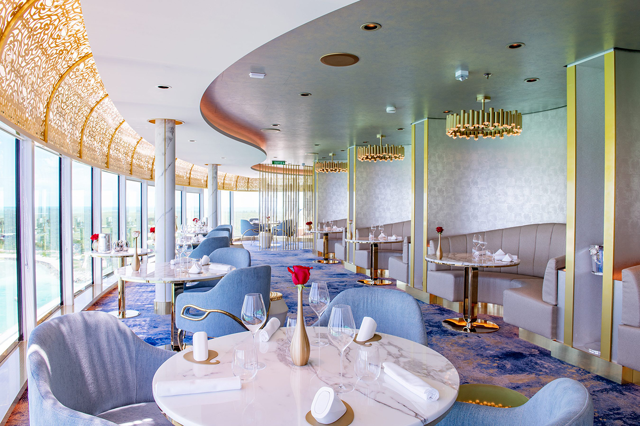 A modern, upscale restaurant with blue and gold decor, featuring curved windows with outdoor views, tables set with white linens and glassware, plush seating lining the walls and floor – an exquisite dining experience reminiscent of Disney Cruise Line's signature style.