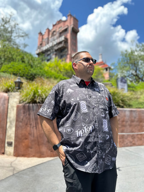A man in a patterned button-up shirt and sunglasses stands outdoors with the Tower of Terror in the background on a sunny day.