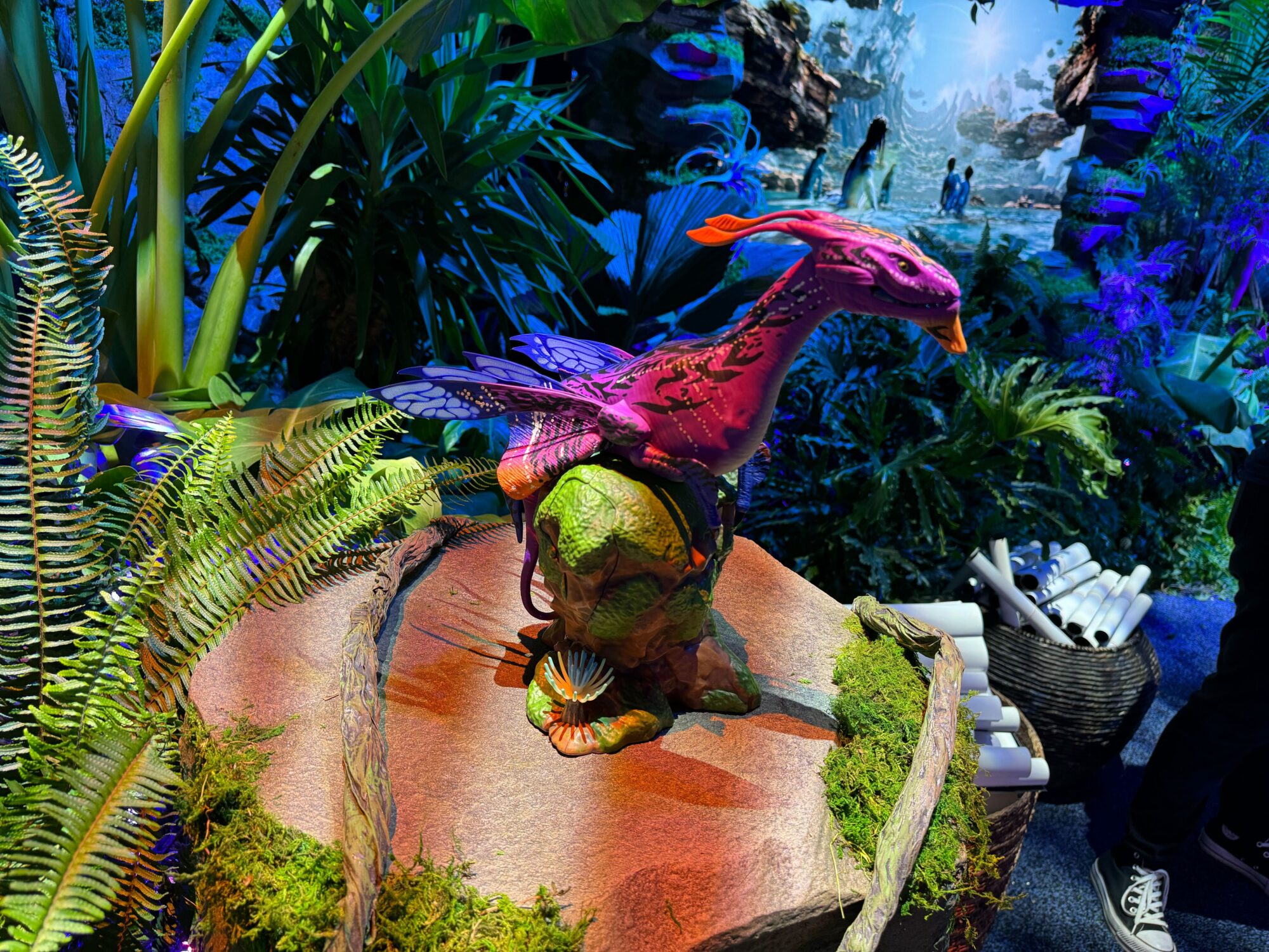A colorful, detailed sculpture of a fantastical creature with wings and a long neck is positioned on a large rock. The background features lush, vibrant vegetation and a person standing nearby.