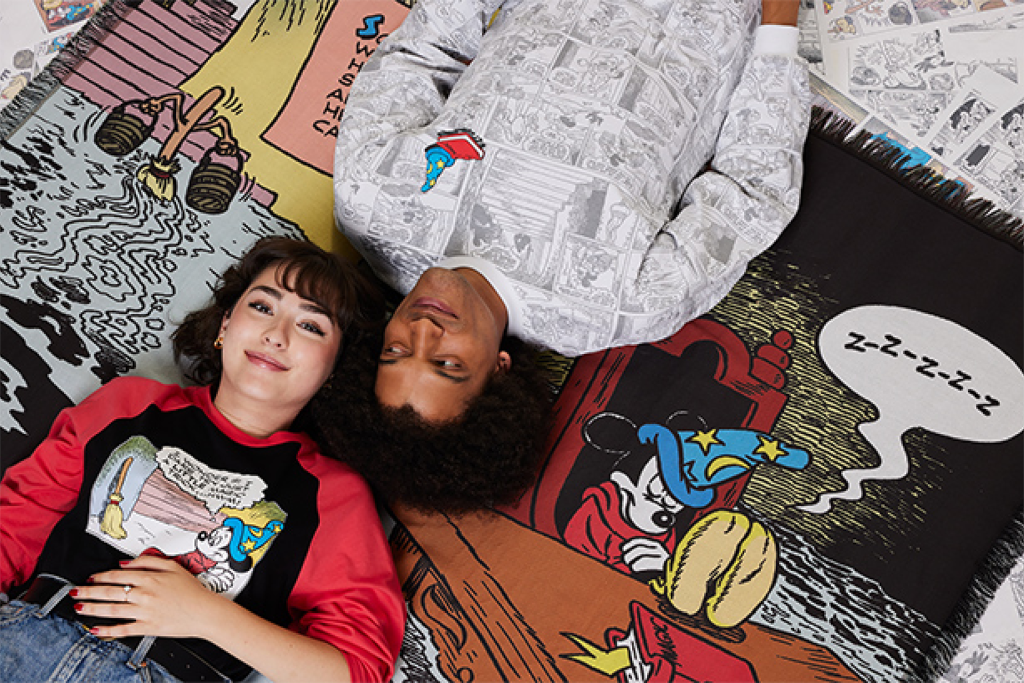 Two individuals lie on a tapestry featuring Mickey Mouse comic strips, with their heads close together. Comic books are scattered around them. Both are wearing graphic tops.