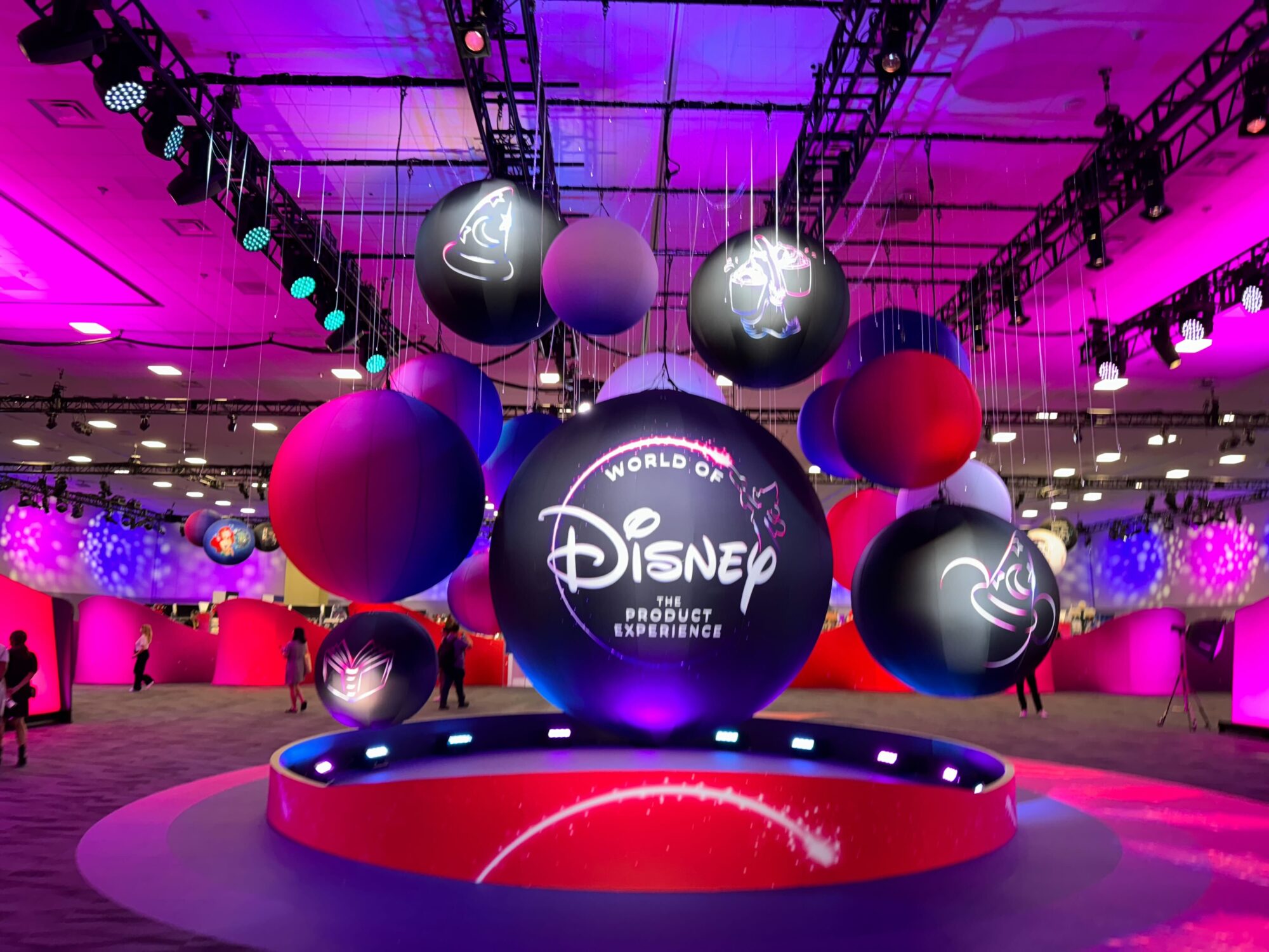 A colorful exhibition display with a central sign reading "World of Disney: The Product Experience," surrounded by hanging spherical decorations and vivid lighting.