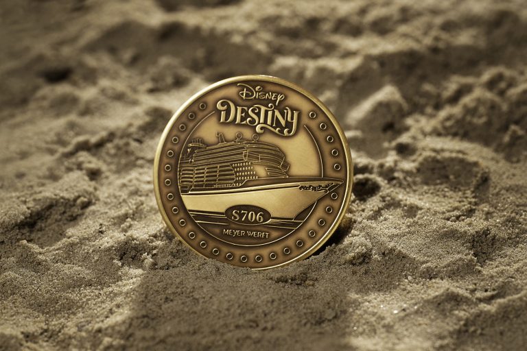 A gold-colored coin featuring a ship with the text "Disney Destiny S706 Meyer Werft" partially embedded in sandy terrain.