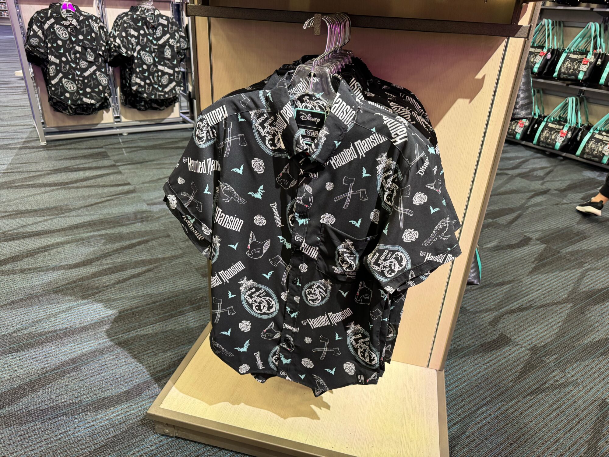 A display of RSVLTS black short-sleeve button-up shirts, celebrating the 55th Anniversary of the Haunted Mansion, featuring various white and light blue graphics and text, hanging on a rack in a store.