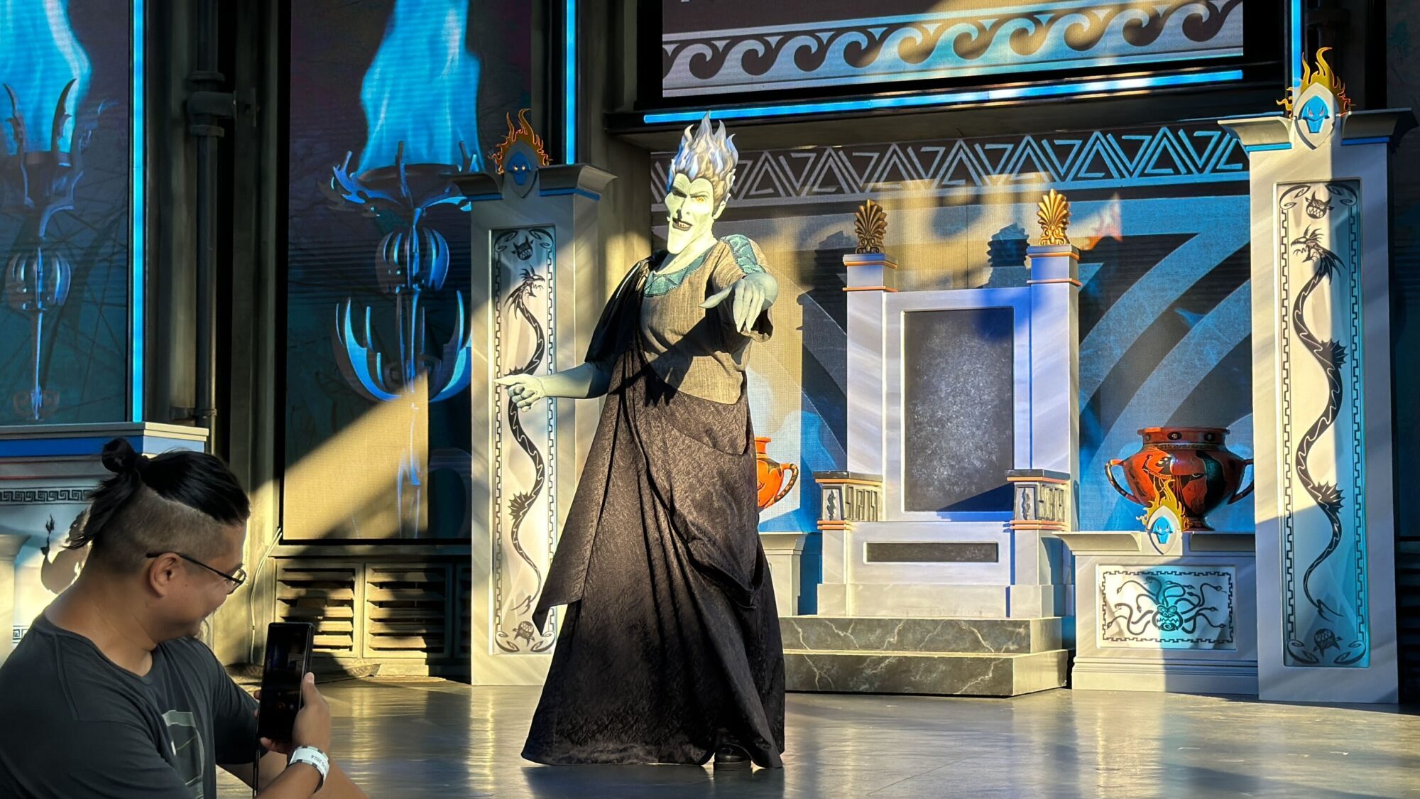 At the Oogie Boogie Bash 2024, a person dressed as a character with green skin and white hair performs on a stage decorated with a throne and columns, looking like Hades himself, while an audience member records the mesmerizing performance with a smartphone.