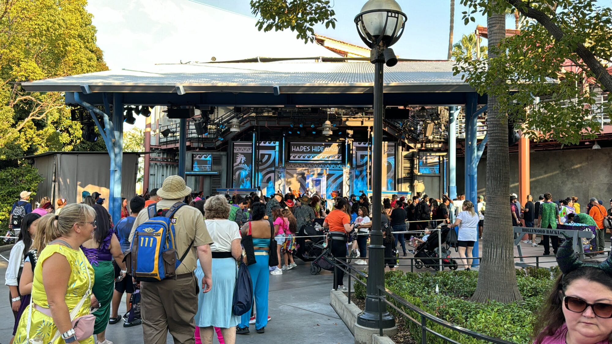 A crowded outdoor stage area with people gathered in the foreground. A sign reading "Hades Hangout" dominates the stage, hinting at an exciting 2024 event. Trees encircle the area, and a lamp post stands to the right, creating an eerie ambiance perfect for Oogie Boogie Bash.