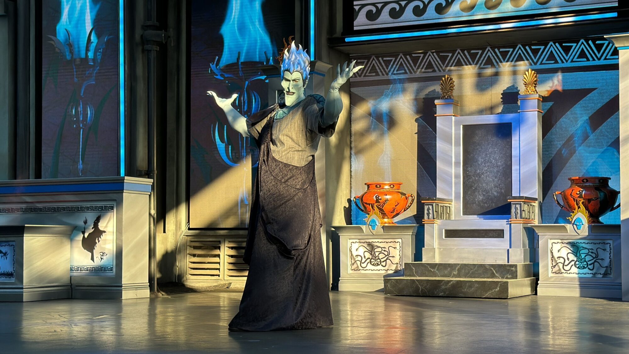 A performer dressed as a blue-haired character with a grayish face stands on a stage at the Oogie Boogie Bash 2024, arms raised. The stage features ornate decorations, including large vases and a patterned backdrop.