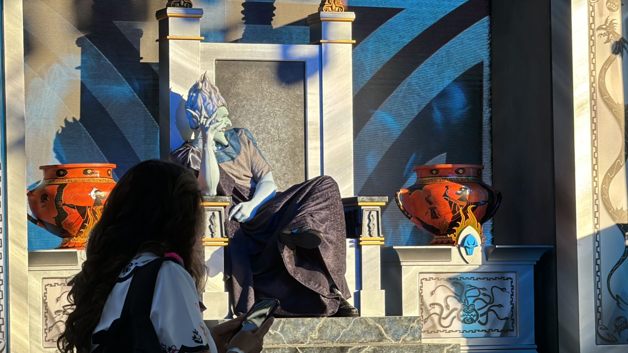 A person in a blue costume and makeup portraying Hades sits on a throne with large vases on either side at the 2024 Oogie Boogie Bash. Another person is seen in the foreground, looking at their phone.