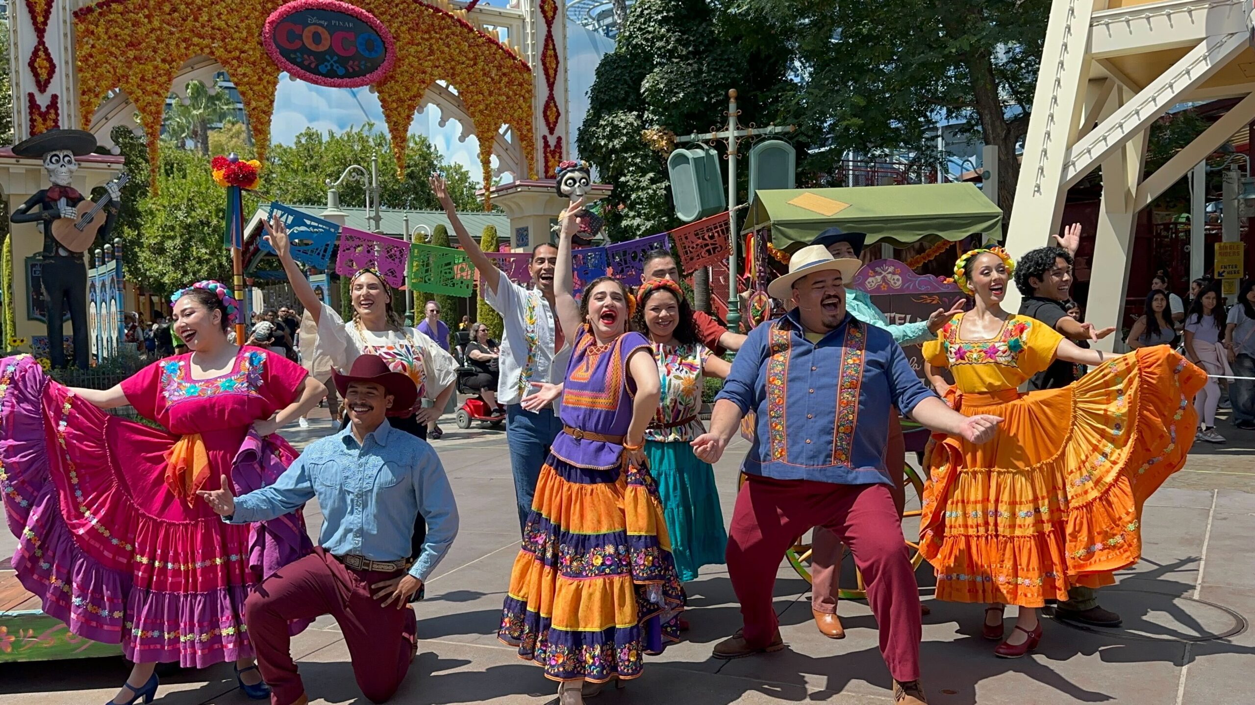 A group of performers in colorful traditional Mexican attire dance and sing in an outdoor setting with festive decorations and a crowd watching.