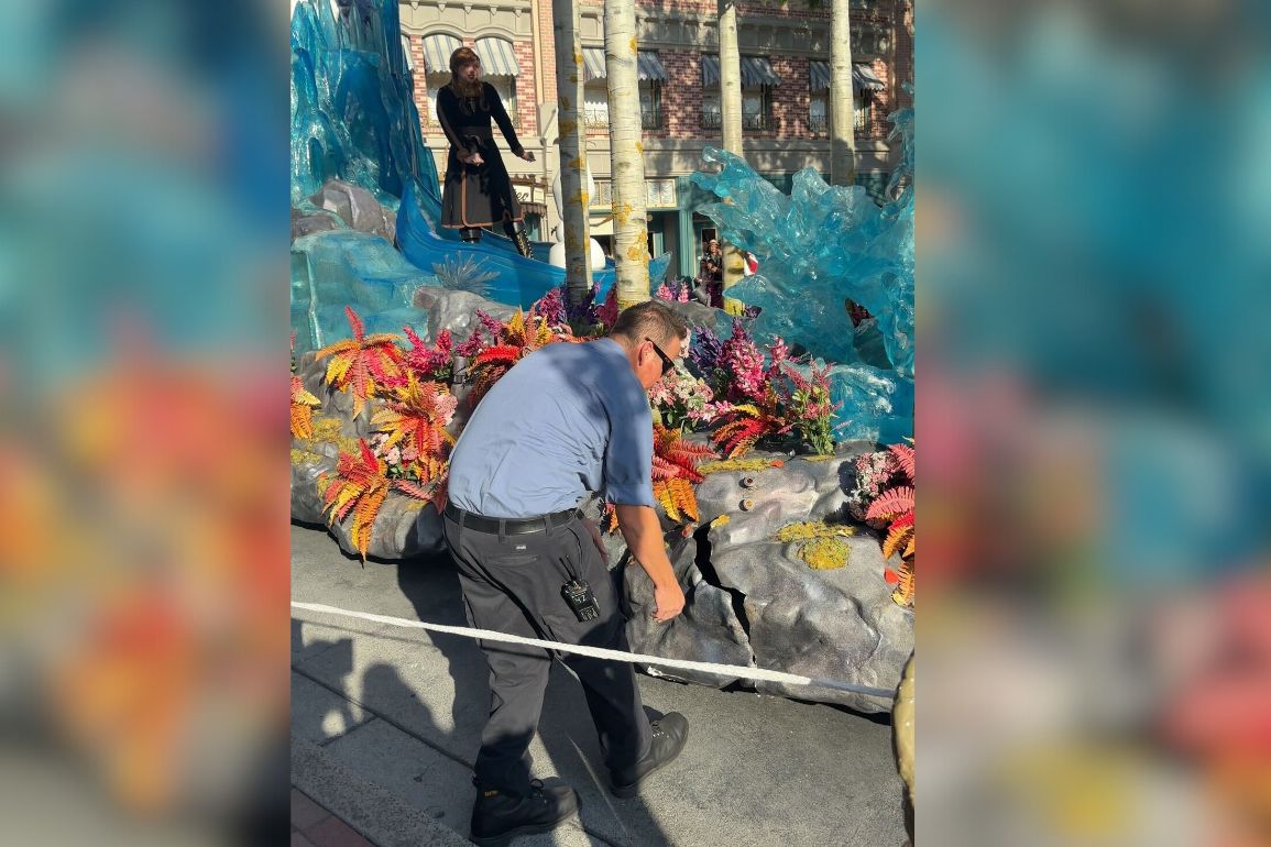 A person in a blue uniform ties a rope around a colorful float decorated with flowers and faux rock formations in an outdoor setting, making sure everything is secure to prevent any float crashes. Another person stands on the float in the background.