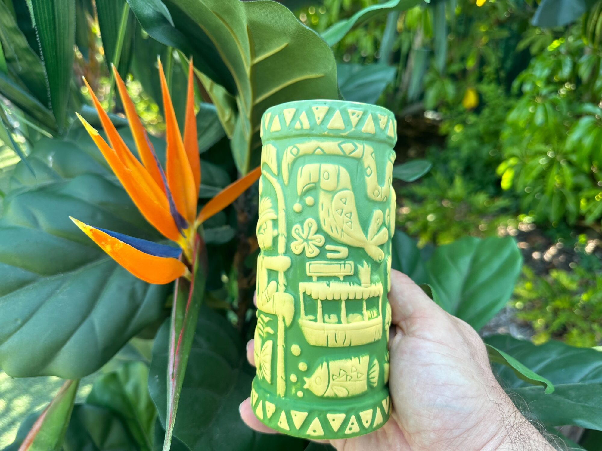 A hand holds a green Skipper's Adventure tiki mug adorned with carvings of tropical scenes and animals, in front of large green leaves and an orange bird of paradise flower.