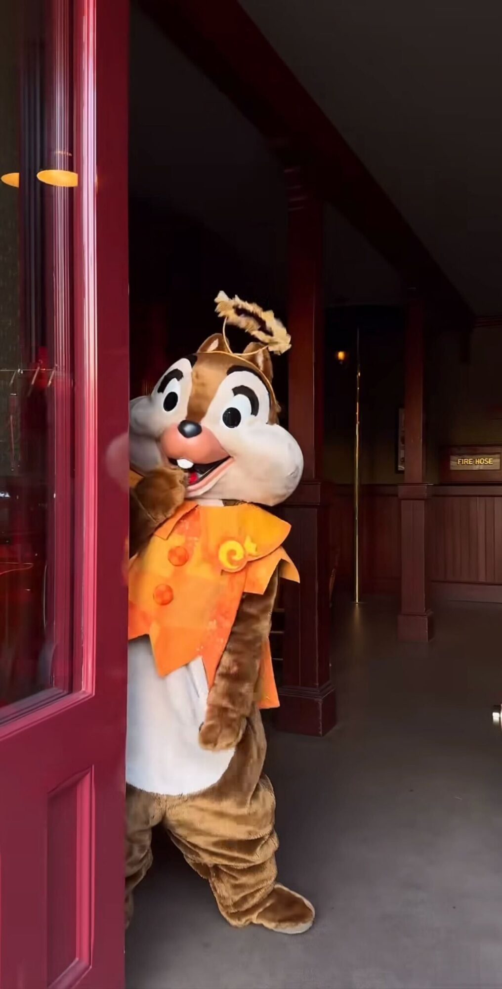 A person in a chipmunk costume stands partially through a red door, inside a dimly lit room with wooden walls.