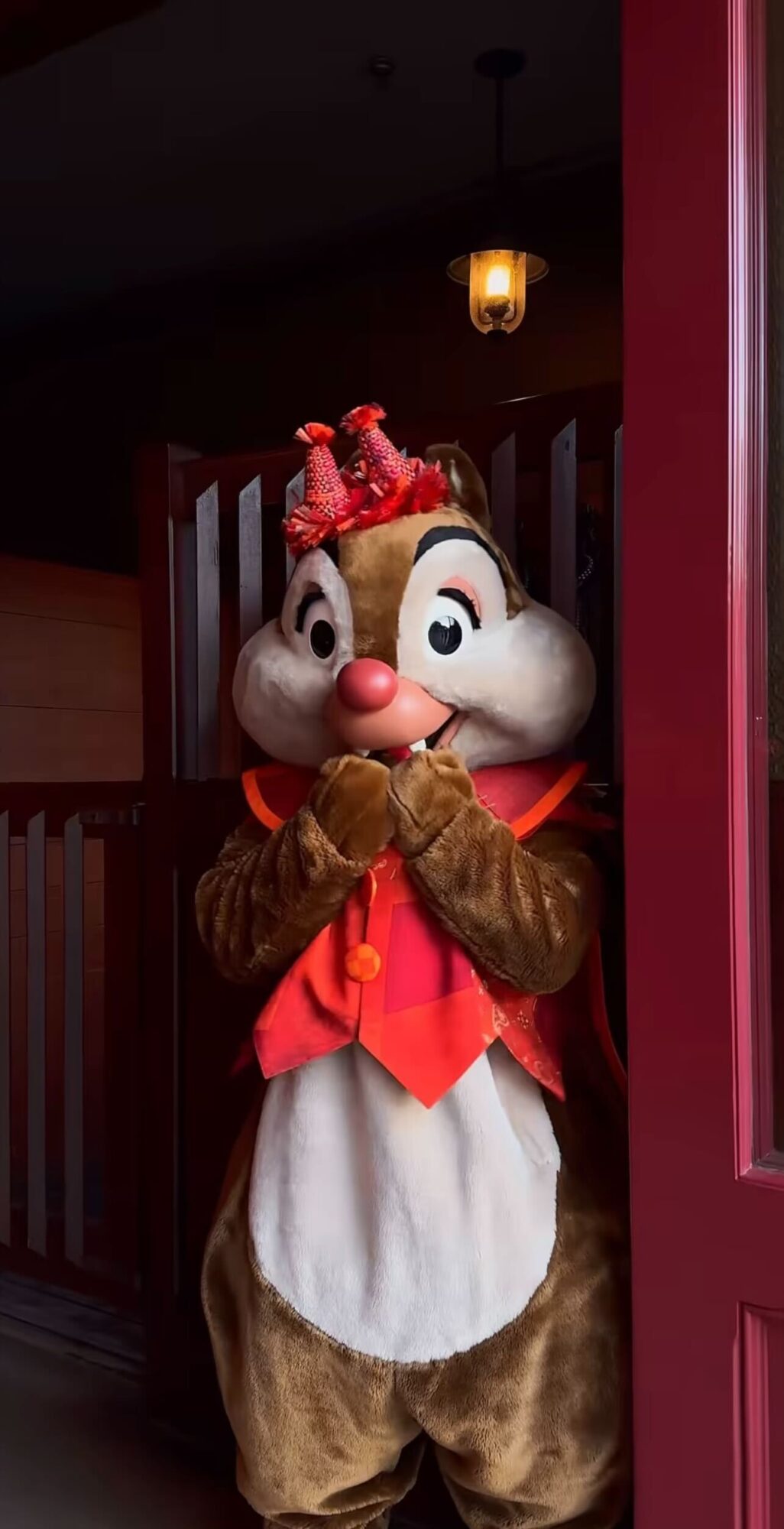 A character in a chipmunk costume with a red outfit and a matching hat stands in a doorway, hands clasped together. A light fixture is visible in the background.