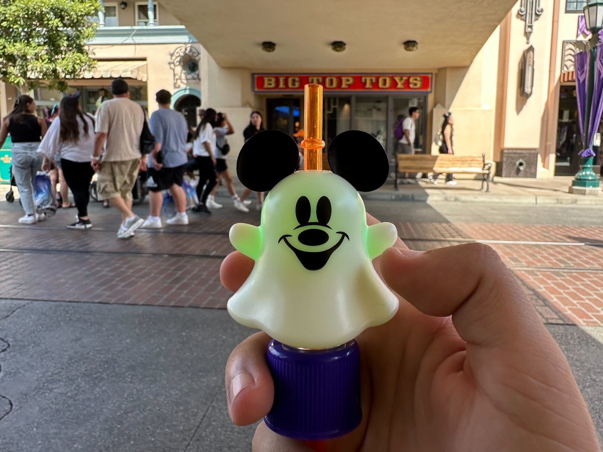 A hand holds a Mickey Mouse-shaped ghost light-up toy, doubling as a Ghost Mickey Bottle Topper, in front of a toy store named "Big Top Toys," while people walk by in the background.