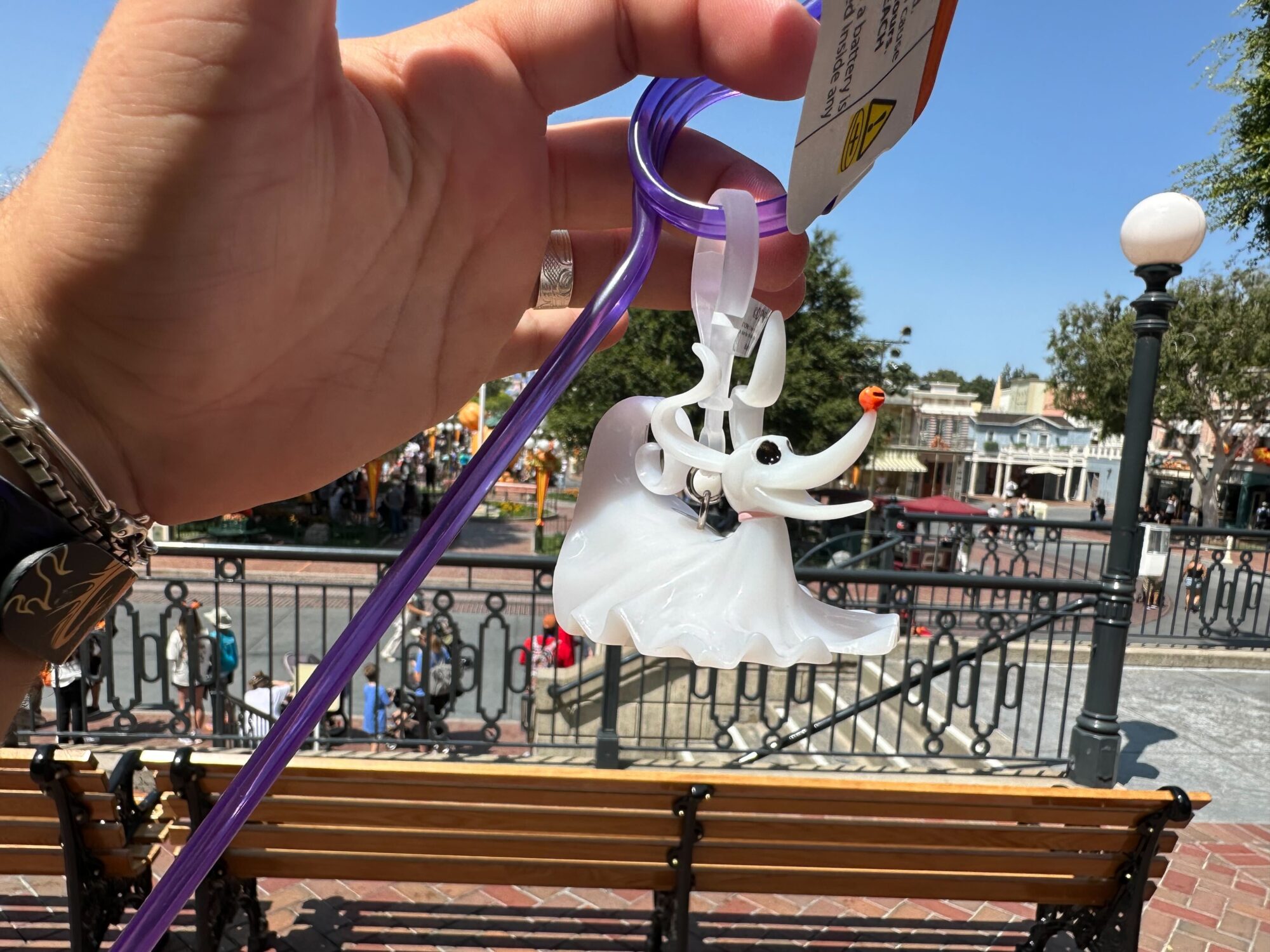 A person holds up a translucent white ghost keychain with a purple string outdoors, in front of a park bench and a railing, resembling Zero from "The Nightmare Before Christmas.