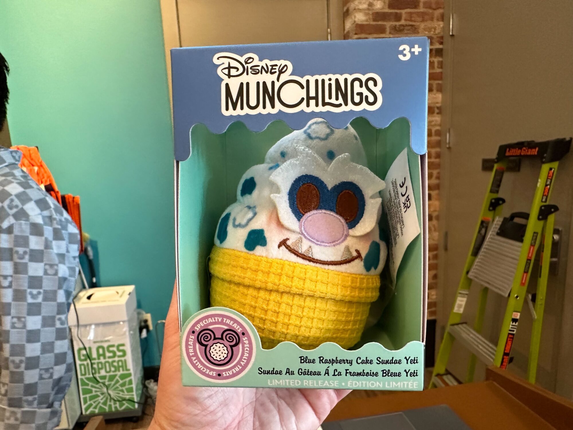 A person holds a box containing a Disney Munchlings plush toy resembling a Blue Raspberry Cake Sundae Yeti.