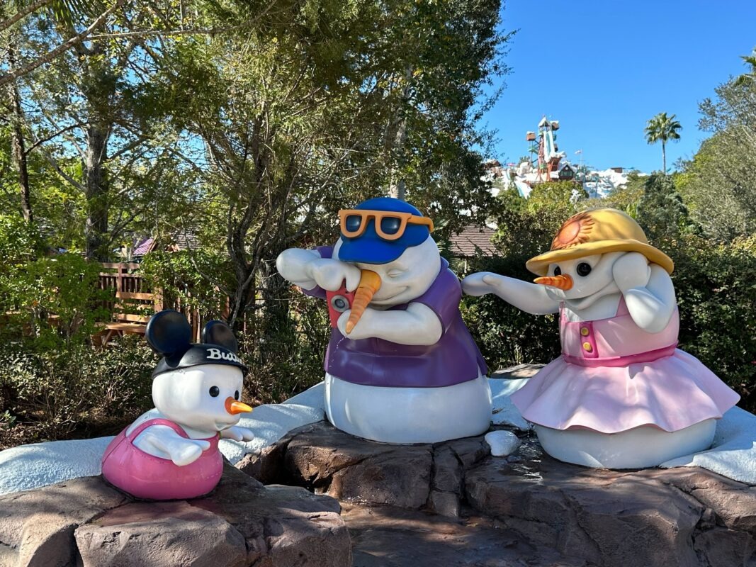Three snowman statues in a park: one wears baby attire and a Mickey hat, another in sunglasses and a jersey with a carrot nose, and a third in a sunhat and dress taking a photo. Trees and attractions create the perfect backdrop for this whimsical daily recap of winter fun.