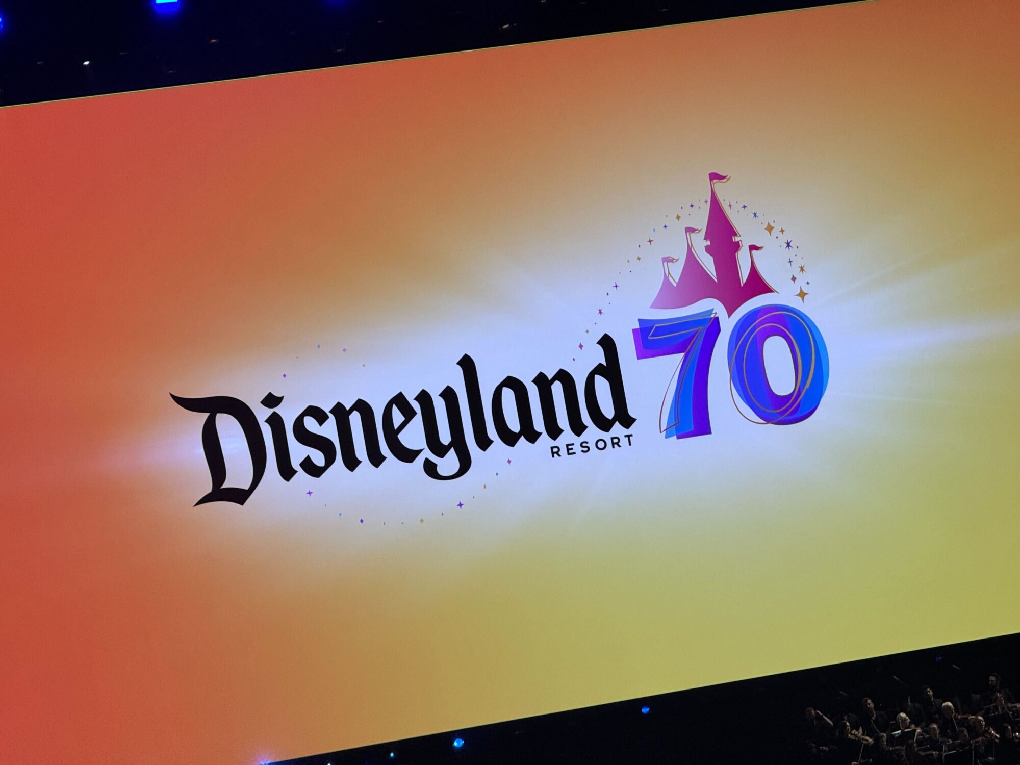 A screen proudly displays the Disneyland logo alongside the number 70, celebrating its grand 70th anniversary. The background is a radiant gradient of orange.
