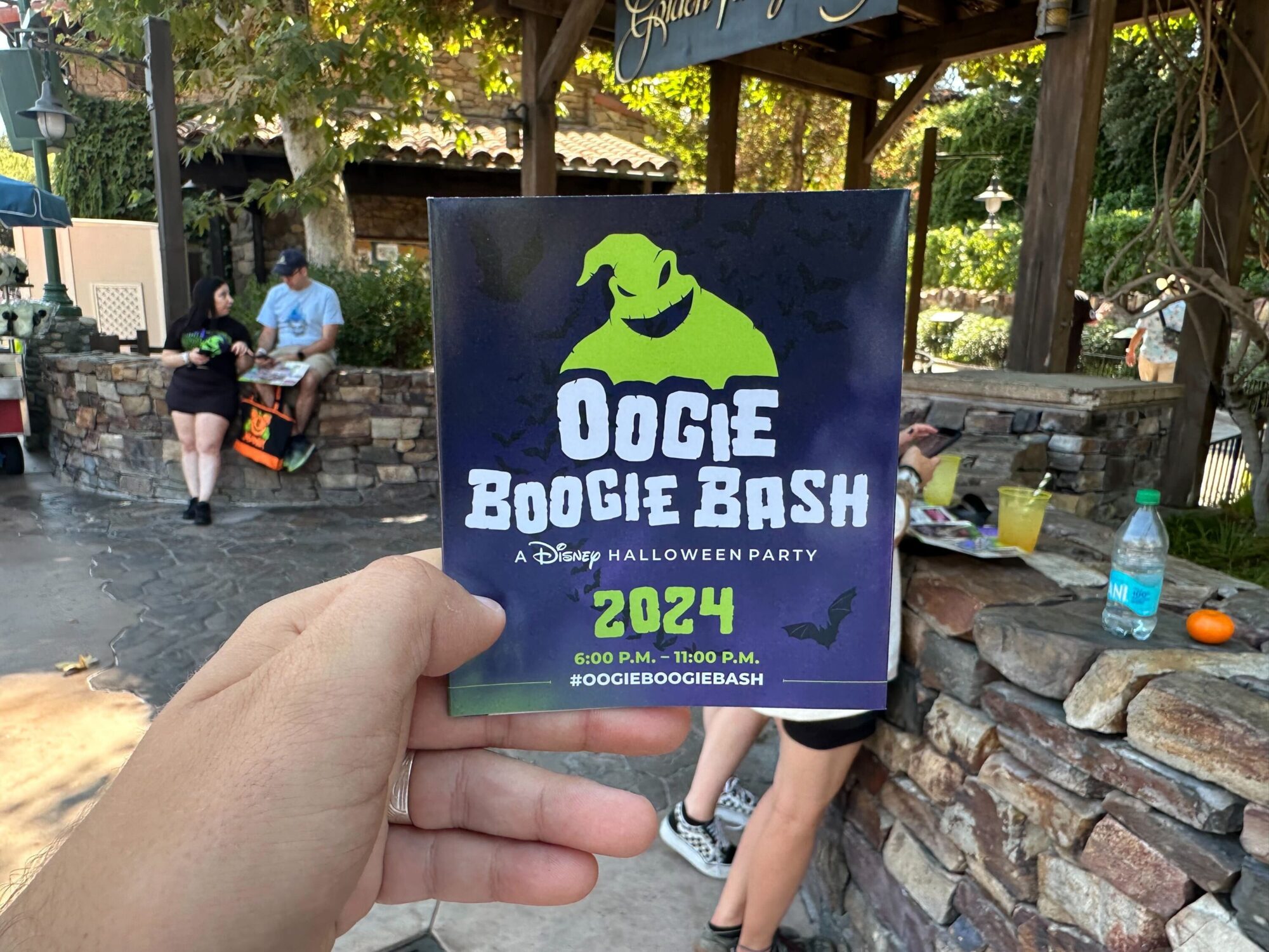 A hand holds a map and flyer for the 2024 Oogie Boogie Bash Halloween party at Disney, with people seated and standing in the background.