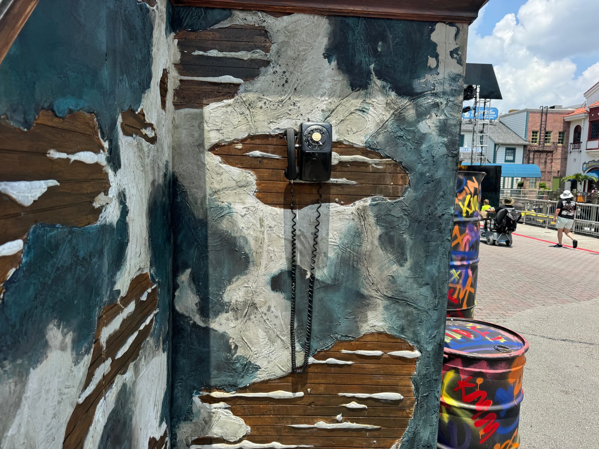 A vintage rotary dial telephone is mounted on a distressed, peeling wall. In the background, a street scene with people, a colorful barrel, and buildings is visible under a partly cloudy sky, reminiscent of the eerie ambiance at Halloween Horror Nights.