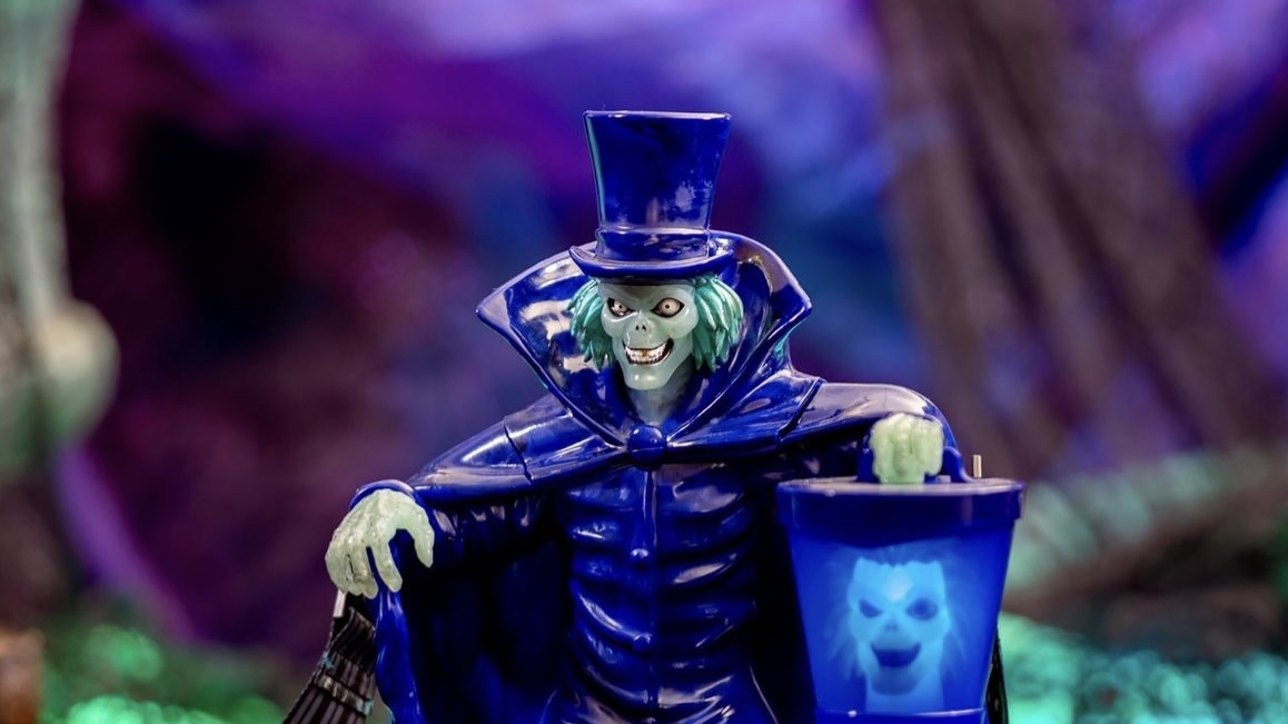 A toy figure is dressed in a blue suit and top hat, holding a small blue bucket with a ghostly face design. The background is a blurry mix of purple and blue hues.