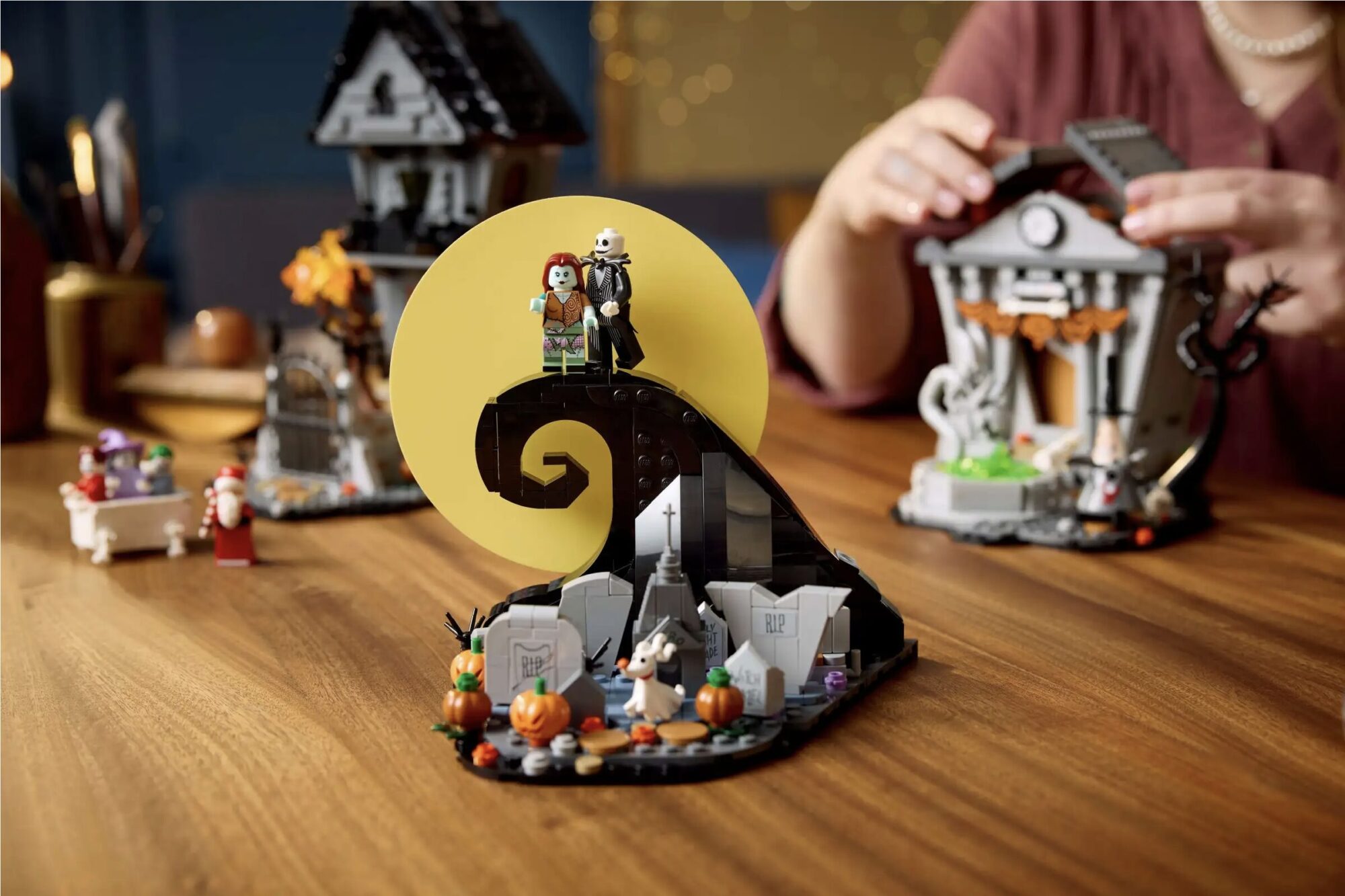 A person is assembling a spooky-themed LEGO set inspired by Tim Burton's "Nightmare Before Christmas," featuring characters and scenes with pumpkins, gravestones, and a curled hill under a yellow moon.