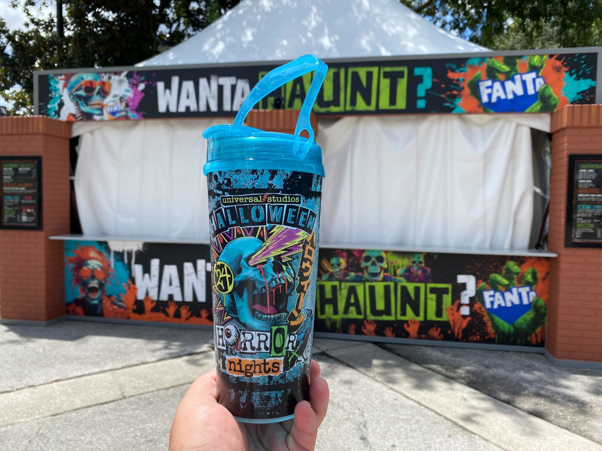 A hand holding a Halloween-themed tumbler with vibrant designs. In the background, there is a colorful "Want a Haunt?" booth with graffiti art and Fanta branding.