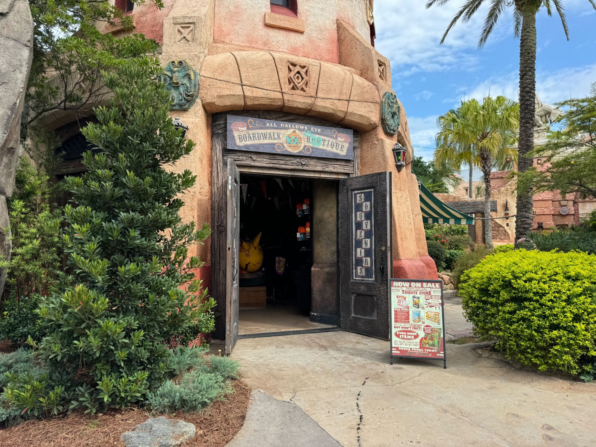 Entrance of a themed store titled "Boardwalk Tattoos" with an open door, inviting interior decor, and colorful signage. Lush greenery surrounds the store, and a stand with promotional information is at the entrance. Entrance of a themed store titled "Boardwalk Tattoos" with an open door, inviting interior decor, and colorful signage. Lush greenery surrounds the store, and a stand with promotional information is at the entrance.