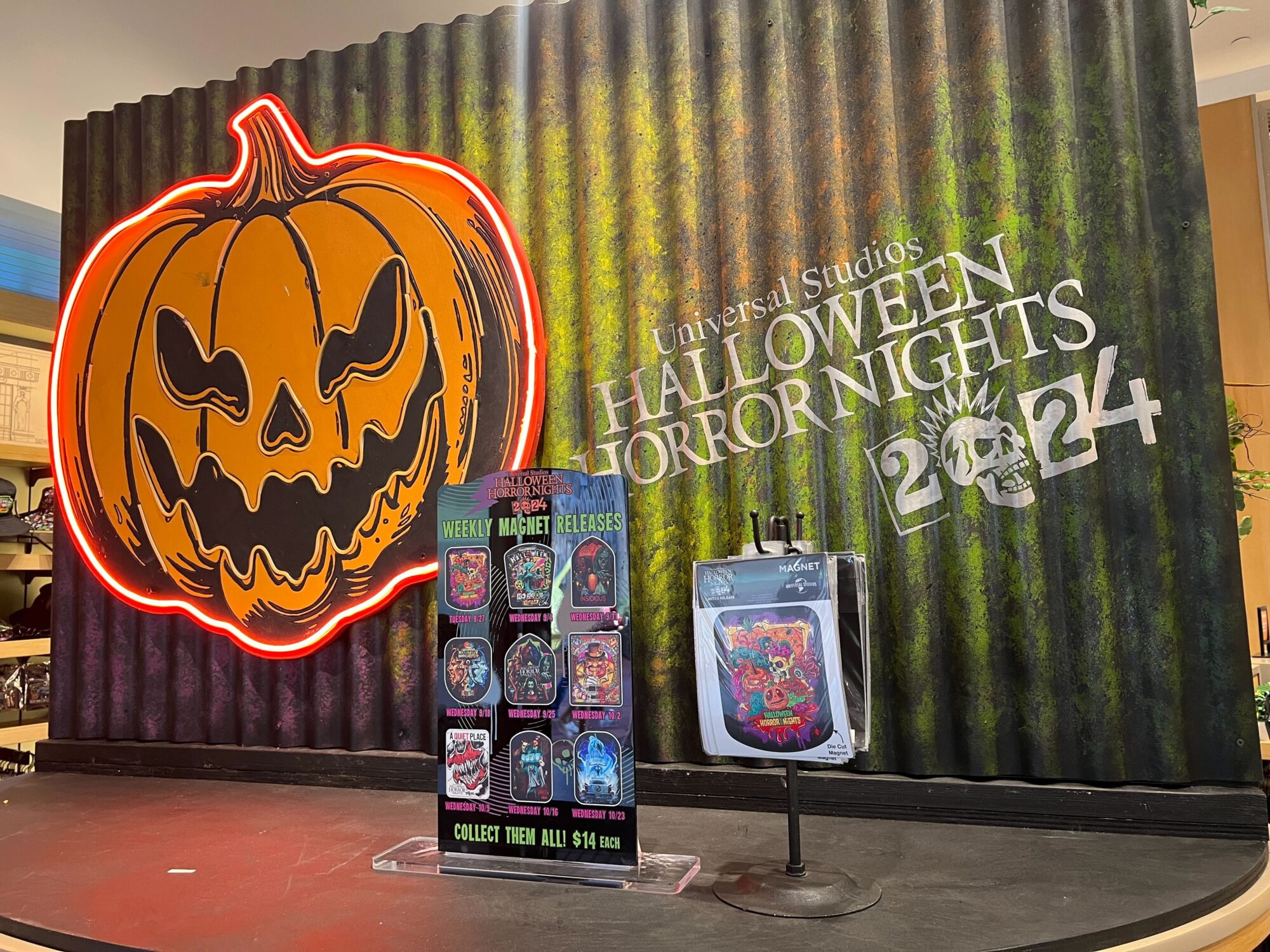 A Halloween Horror Nights 2024 display with a large pumpkin illustration, neon sign, and collectible pin advertisement. Various pins are showcased on a stand in front of the display.