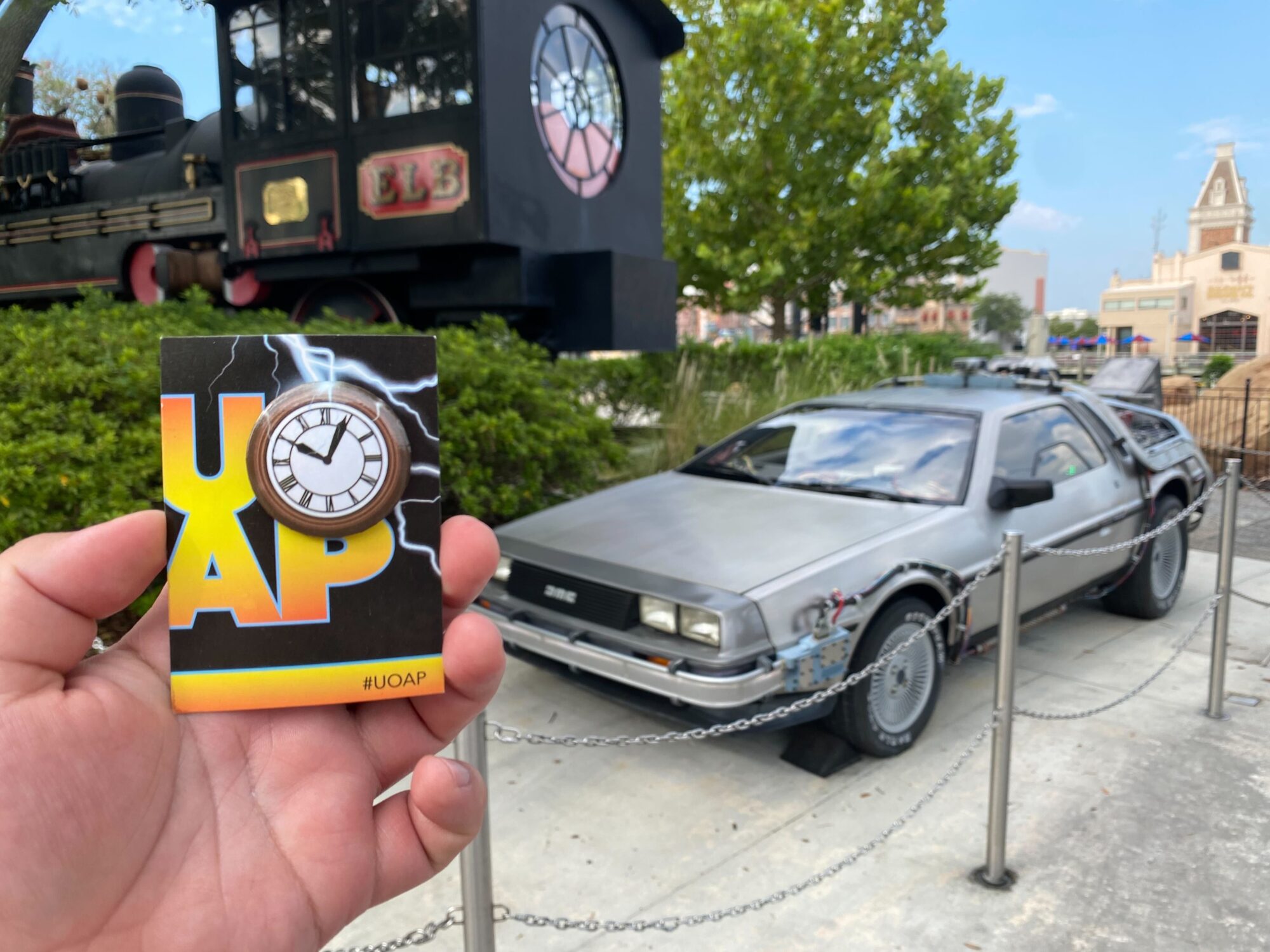 A person holds a "UOAP" badge with a clock design in front of a display featuring a DeLorean car and a time machine from a popular movie series.