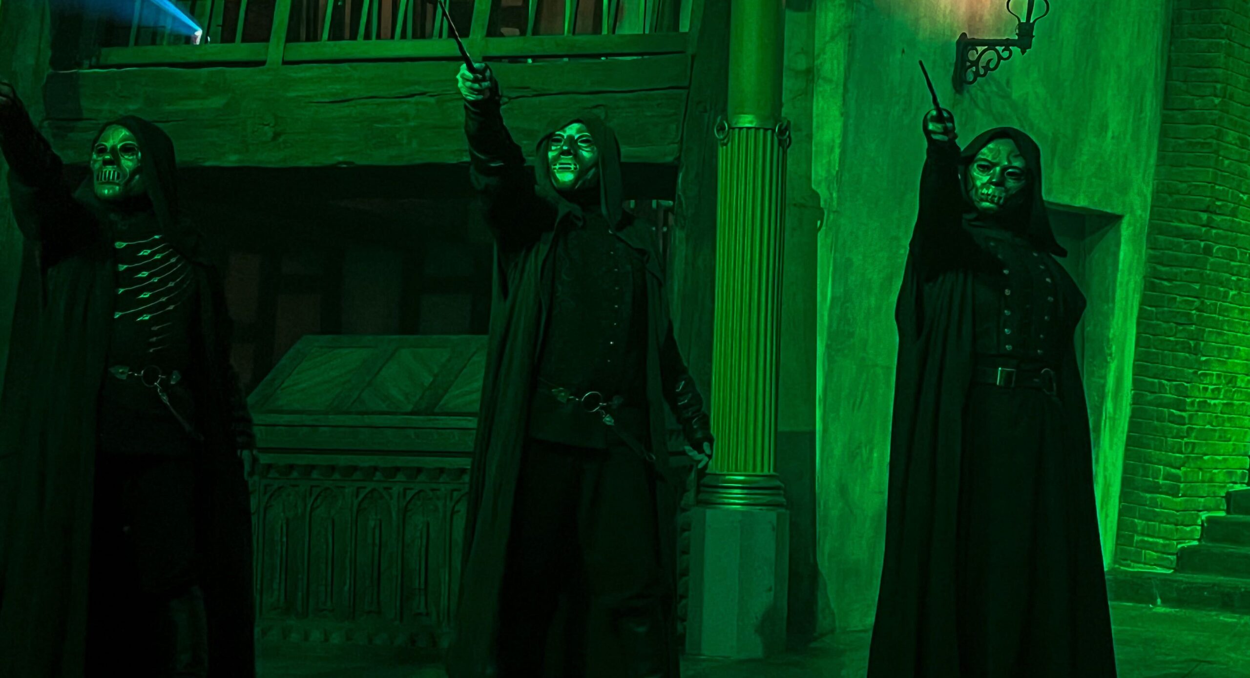 Three people in black robes and masks hold up wands in a dimly lit, green-tinted scene with gothic architecture, reminiscent of an eerie night in Diagon Alley.