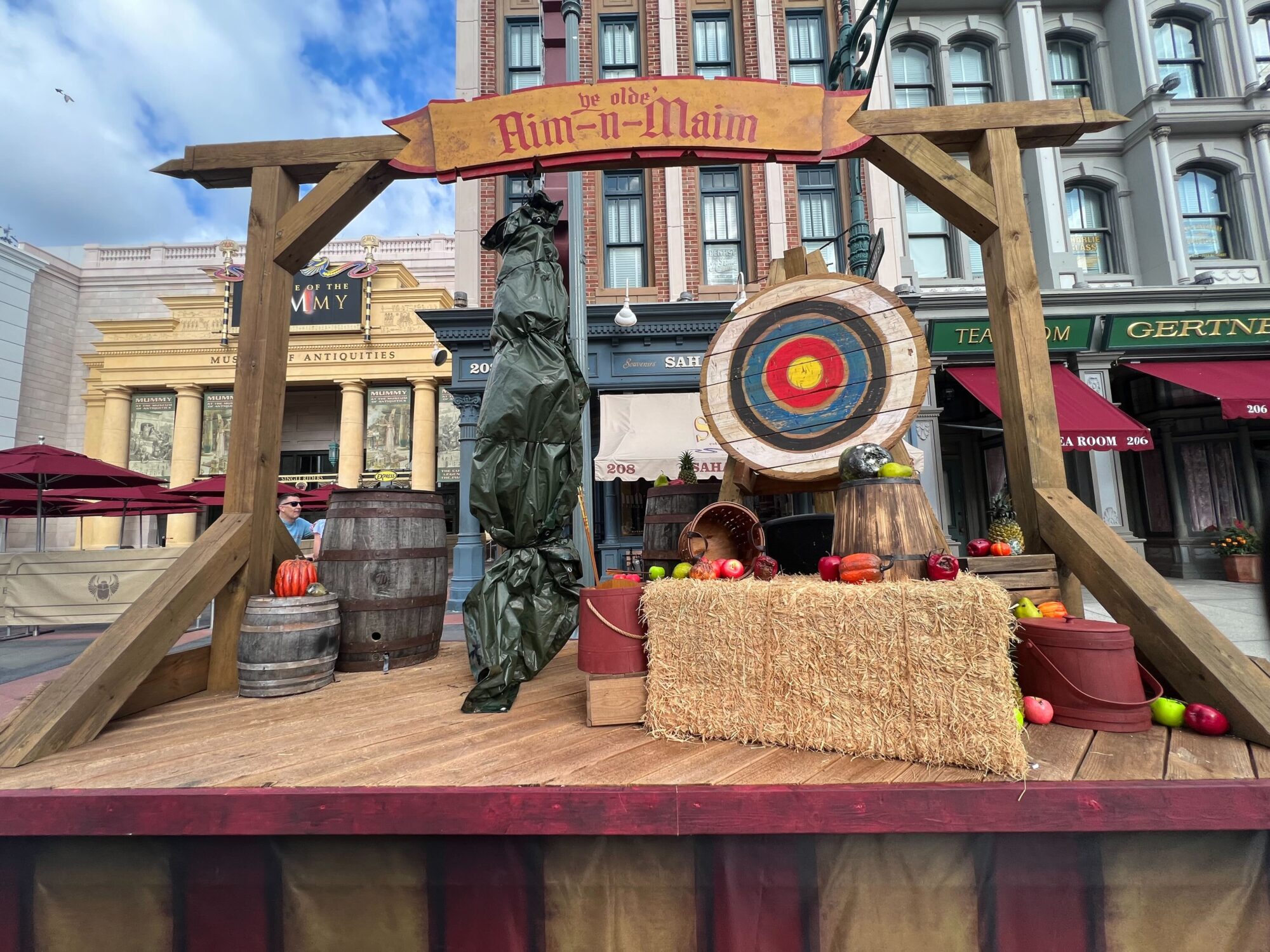 A wooden stage with pumpkin decorations, barrels, a covered figure, and a hay bale with produce, featuring an archery target. Buildings with storefronts are in the background under a partly cloudy sky.
