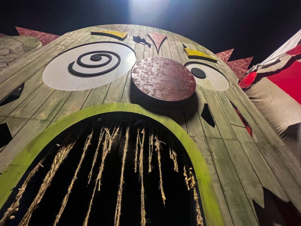 Close-up view of a large wooden carnival funhouse entrance with a painted face featuring exaggerated eyes and a wide-open mouth with ropes hanging down, unmasking the horror that awaits inside.