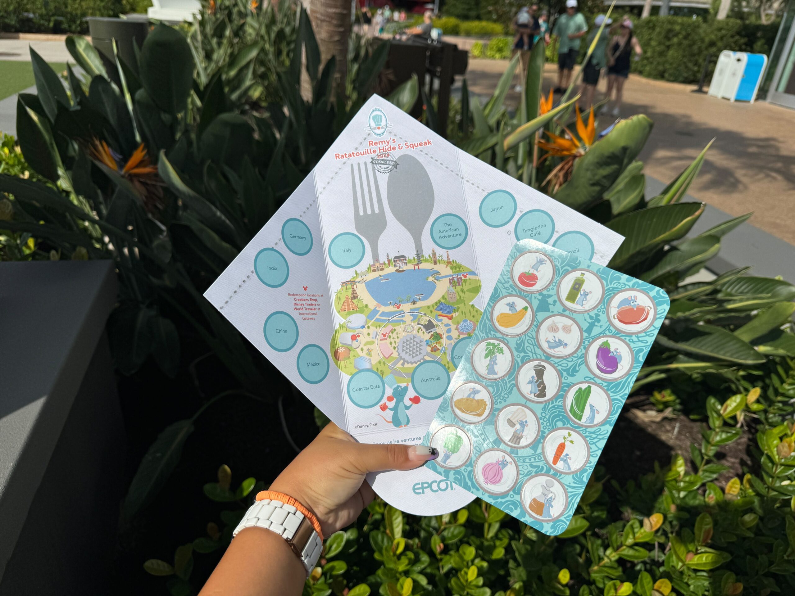 A hand holds a 2024 Scavenger Hunt map and sticker sheet in front of a garden area at Remy's Hide & Squeak during the EPCOT Food & Wine Festival.