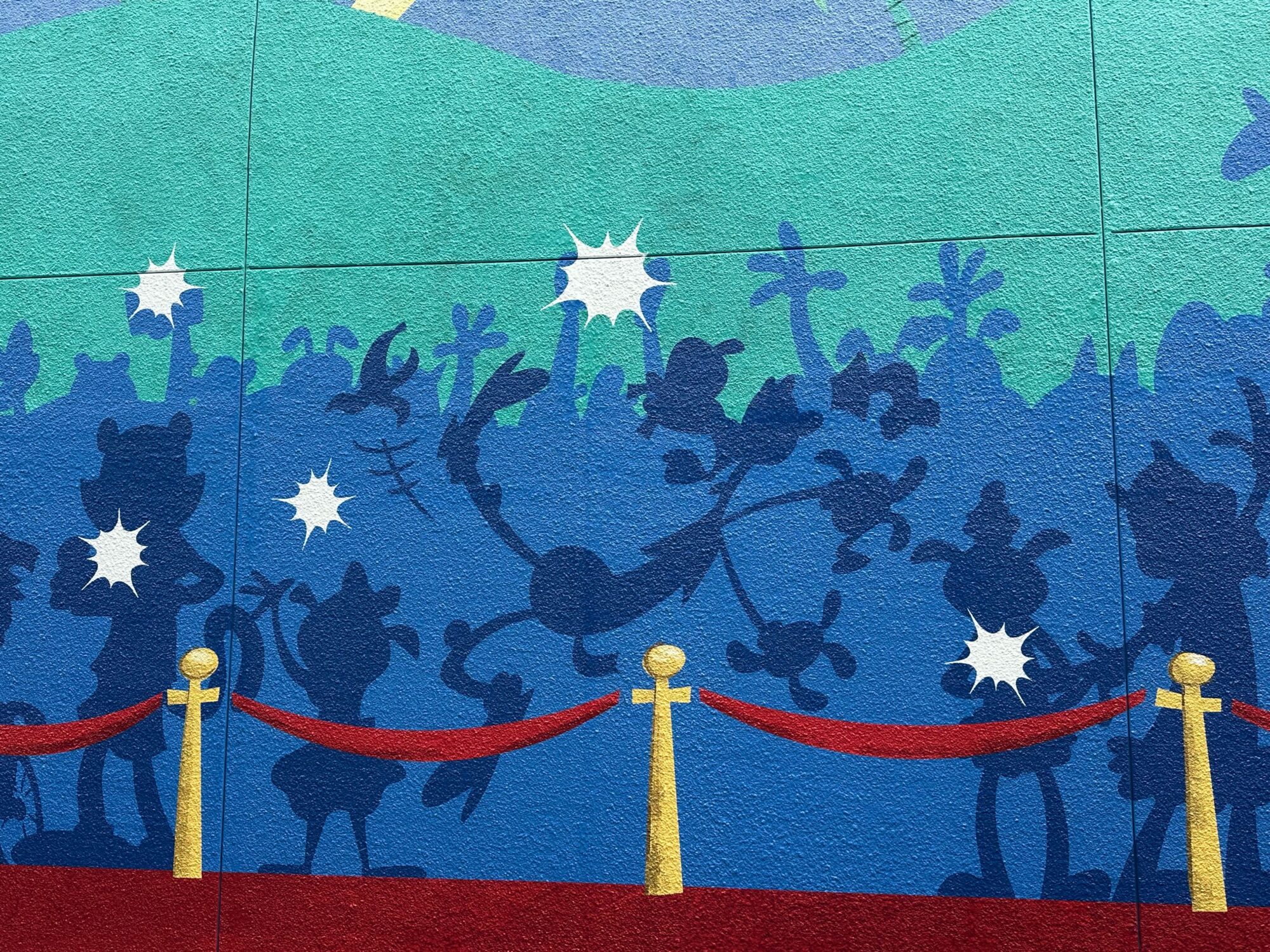 A mural features blue silhouettes of cartoon characters in a lively scene, some striking poses and others swinging. Red ropes on gold posts create a barrier in front of the artwork.